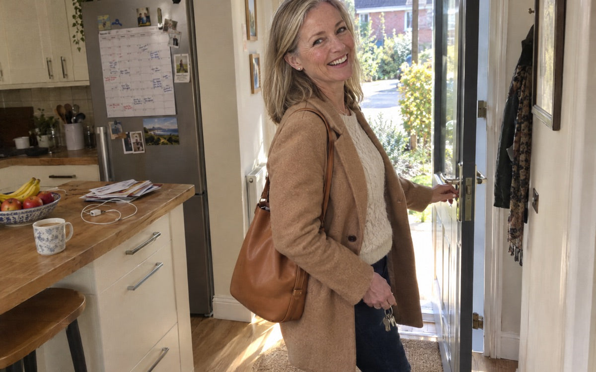 Soft morning light in a quiet kitchen: a woman in her sixties at the window with coffee, dressed to leave the house, no product in frame