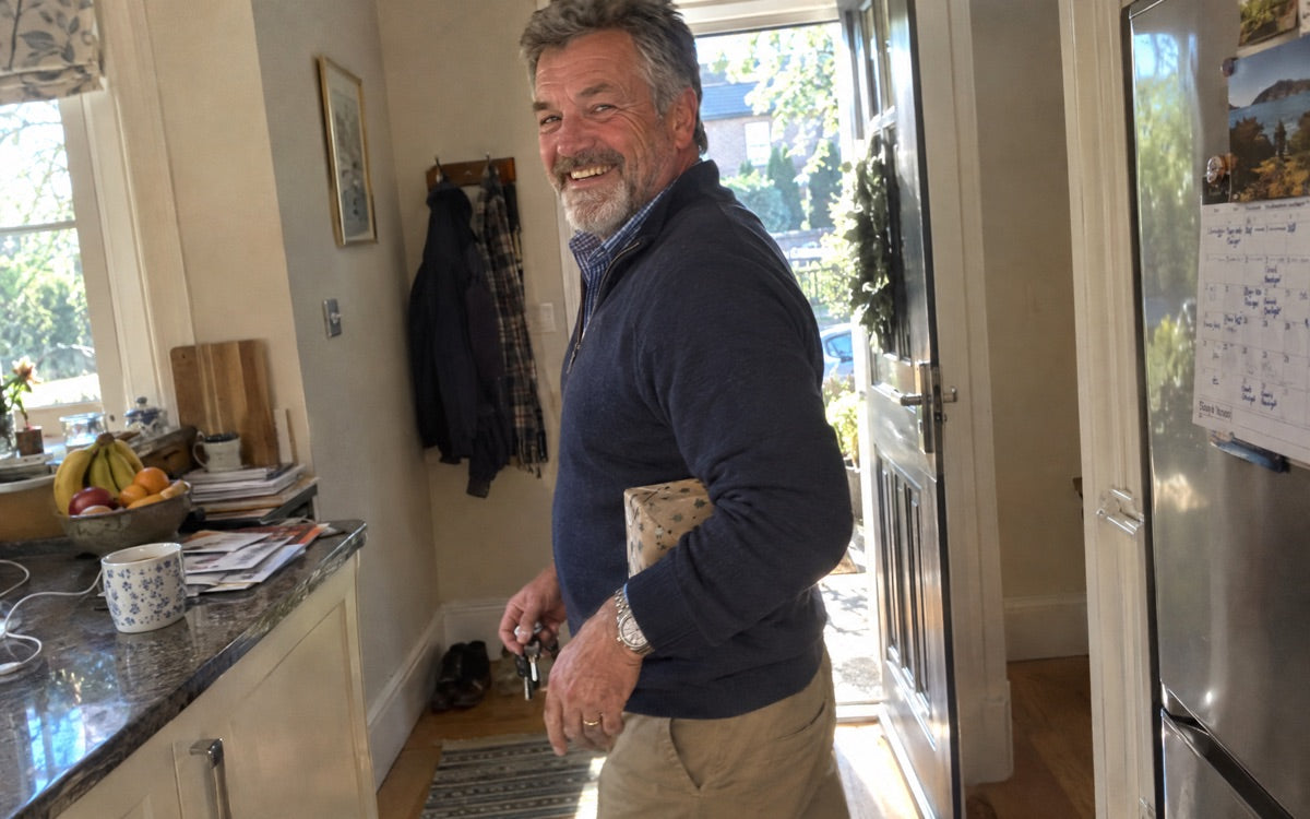 Soft morning light in a quiet kitchen: a man in his sixties at the window with coffee, dressed to leave the house, no product in frame